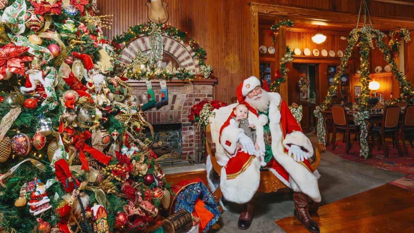 Hire a Santa Claus near me in Atlanta: Santa seated by a fireplace and tree, holding a baby.