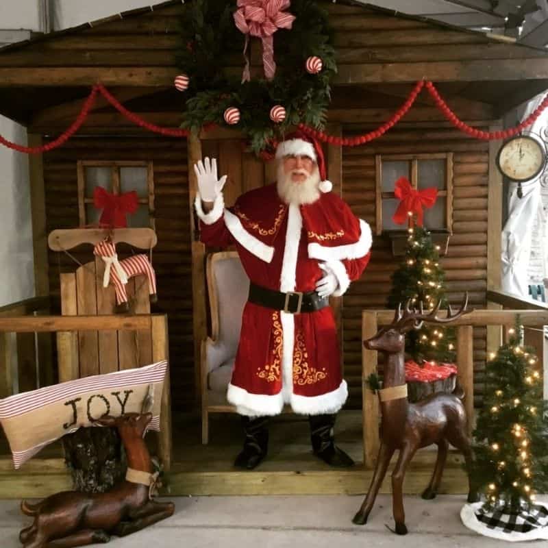 Real bearded Santa for hire waving from a log-cabin set with wooden reindeer.