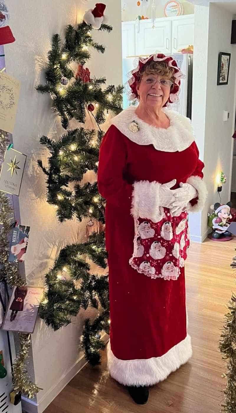 Mrs. Claus for hire in Atlanta standing by a lit tree in a red dress and Santa-print apron.