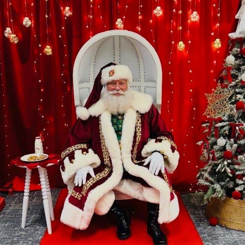 Hire Santa in Atlanta: real beard Santa seated in a white chair with cookies, milk, and a tree.