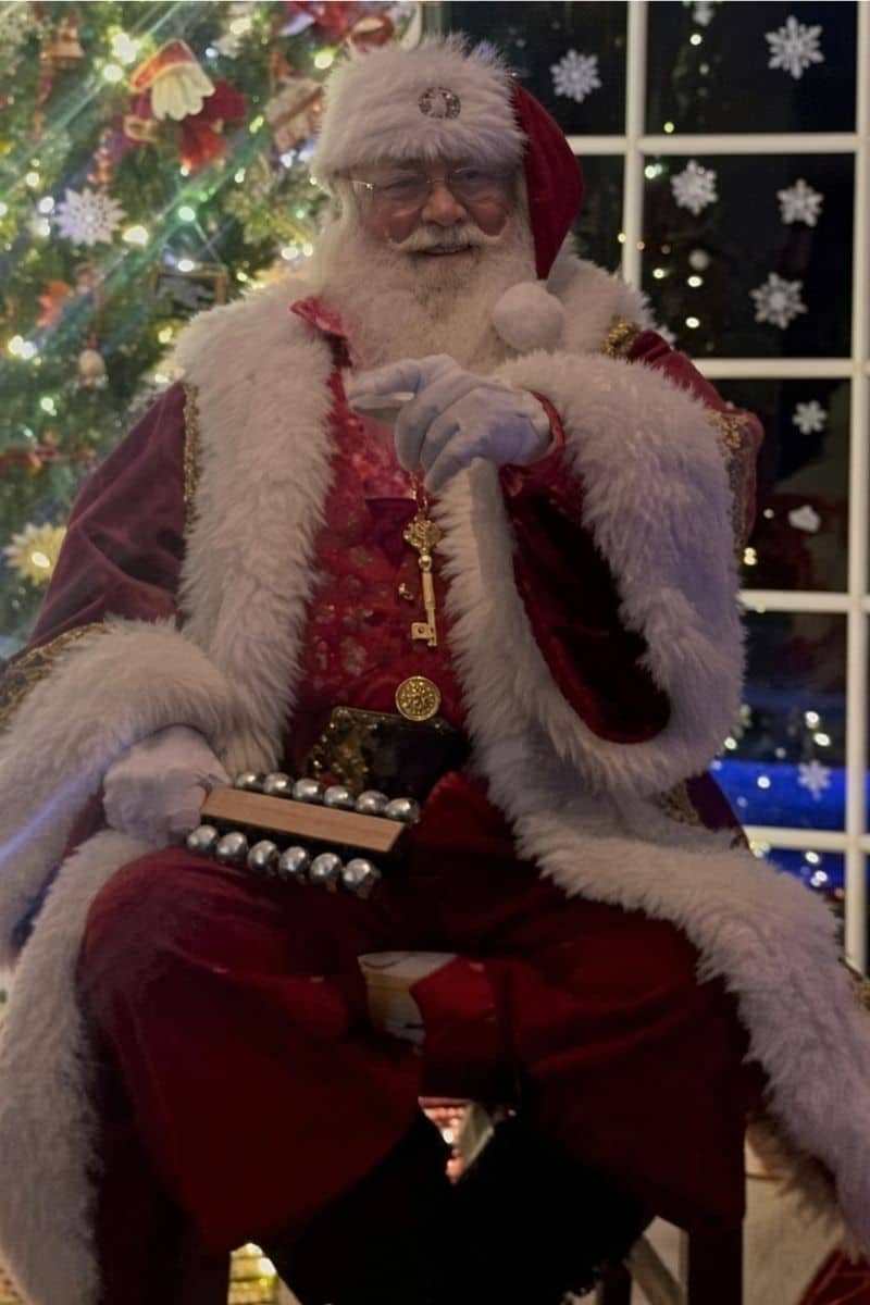 Hire a Santa Claus near me: Santa holds sleigh bells by a tree and a night window in Atlanta.